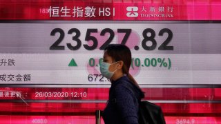 In this March 26, 2020, file photo, a woman wearing face mask walks past a bank electronic board showing the Hong Kong share index at Hong Kong Stock Exchange.