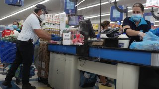 Lay Guzman stands behind a partial protective plastic screen and wears a mask and gloves as she works as a cashier at the Presidente Supermarket on April 13, 2020 in Miami, Florida.