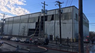 A large warehouse with fallen siding, with firehoses and water on the ground