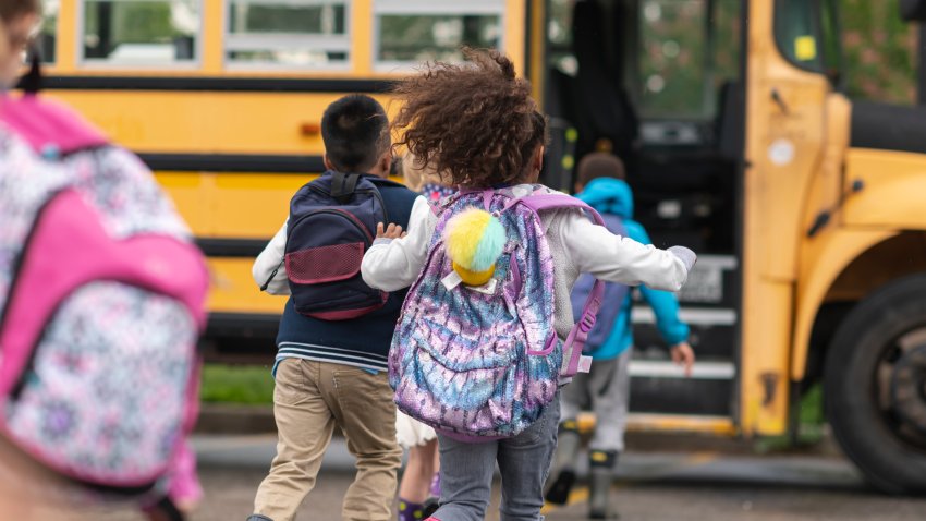 A multi-ethnic group of elementary age children are getting on a school bus. The kids’ backs are to the camera. They are running towards the school bus which is parked with its door open. It’s a rainy day and the kids are wearing jackets, rain boots and backpacks.