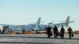 Virgin Galactic pilots walk to the company’s SpaceShipTwo Unity spacecraft, attached to the jet carrier aircraft Eve.