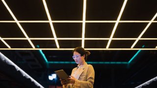 A young Asian business woman wearing glowing smart glasses uses a tablet in a city street at night. Concepts such as augmented reality, artificial intelligence, smart cities, metaverse, post-humanism are expressed.