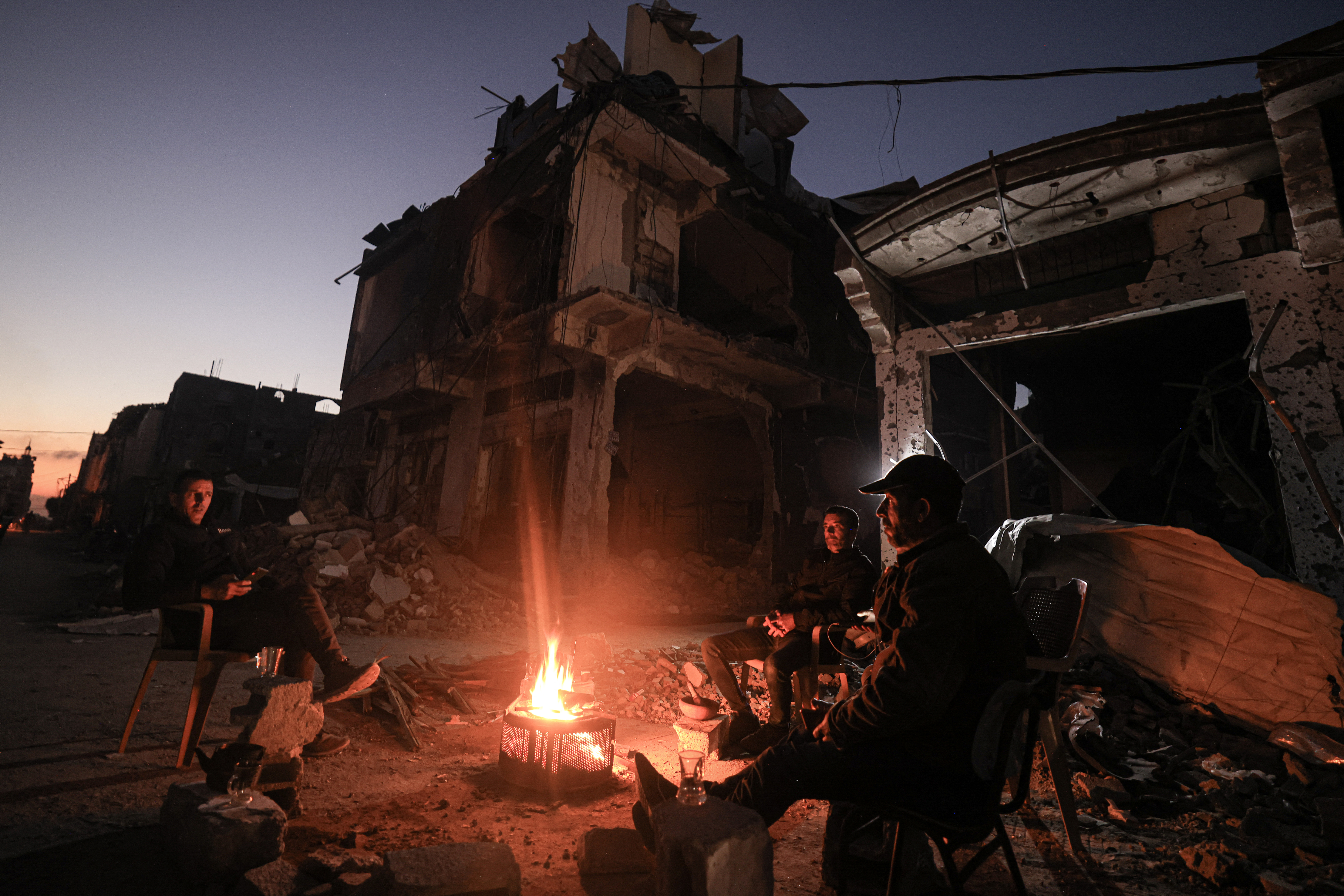 People sit around a camp fire in the midst of destroyed buildings in the Khezaa district on the outskirts of the southern Gaza Strip city of Khan Yunis, following weeks of Israeli bombardment, as a truce between Israel and Hamas entered its 7th day on November 30, 2023.