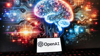The OpenAI logo is displayed on a cell phone with an image on a computer monitor generated by ChatGPT's Dall-E text-to-image model
