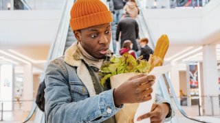 A man with a paper bag of groceries looks surprised and upset at a receipt from a supermarket with high prices against the background of an escalator with customers in the shopping center. 