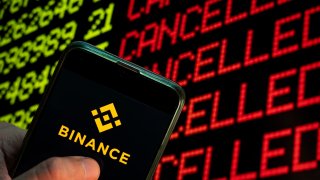 The logo of cryptocurrency exchange Binance displayed on a smartphone with the word “cancelled” on a computer screen in the background.