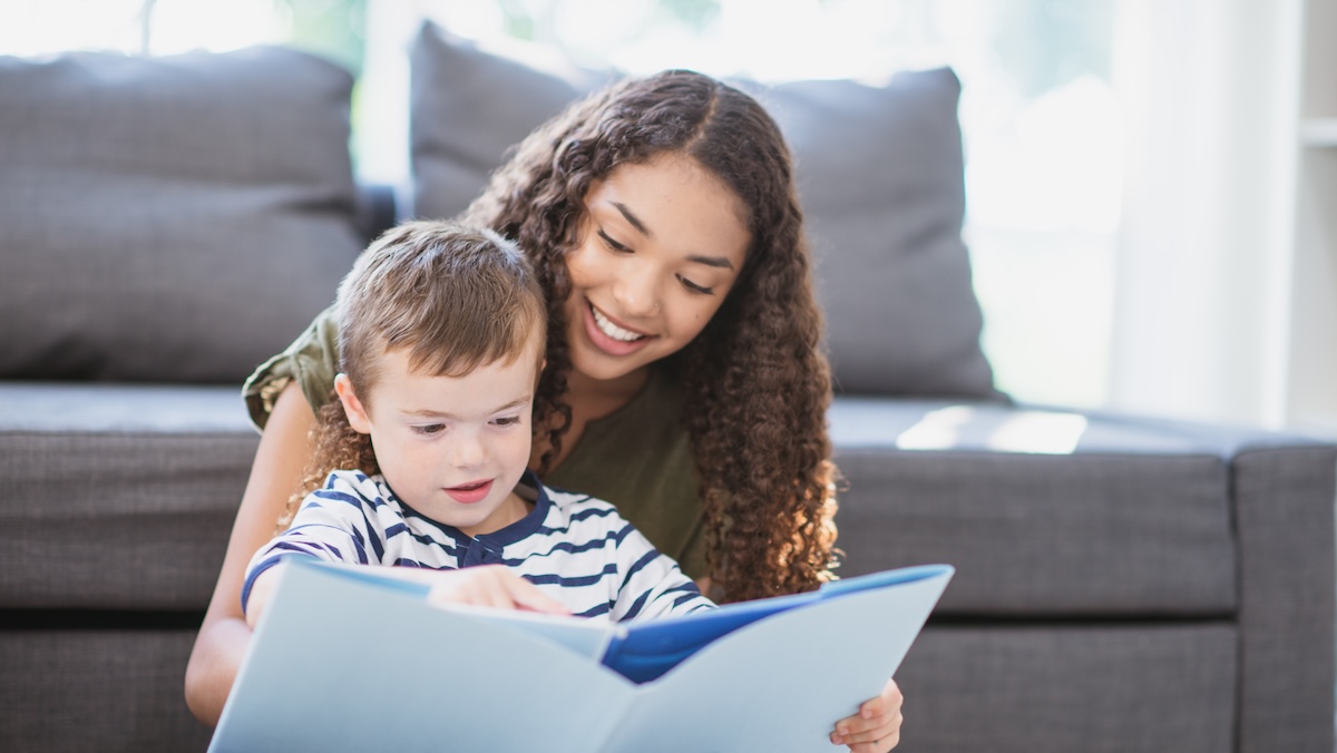 A young boy is with his nanny, a female high school student. They are spending time reading a book together in the living room.